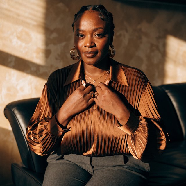Portrait of an adult Black woman in her 50s wearing a pleated copper satin blouse and tailored charcoal trousers, her hands gently holding the blouse fabric, calm half-smile, warm plaster backdrop, late-afternoon window light grazing her face. — Recraft v4 Pro