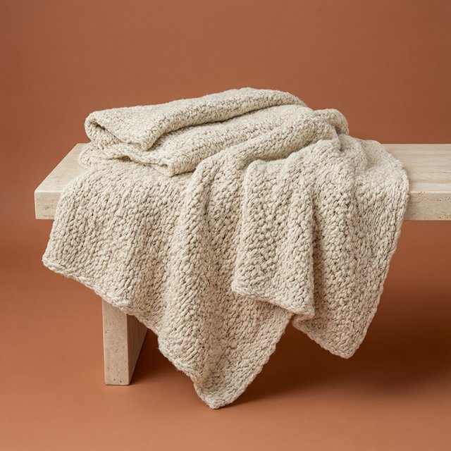 Soft folds of an unbranded chunky-knit wool throw rest on a pale travertine bench; its looped surface catches broad diffused key light and gentle side fill against a muted terracotta studio background, forming a clean commercial product image with no lettering. — Model 18