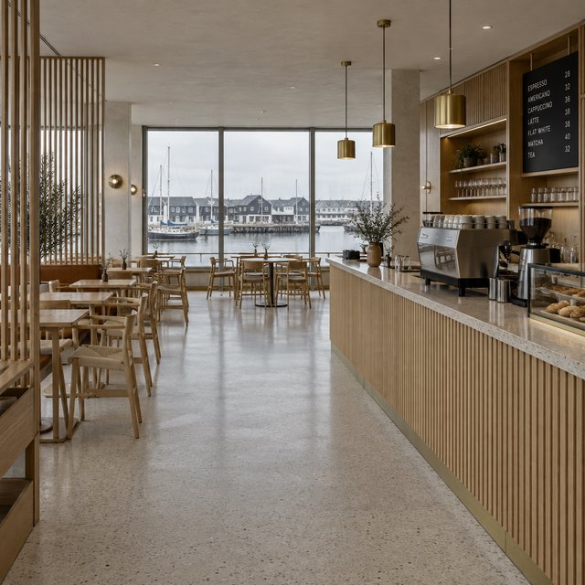 Inside an empty harbor café, restrained Danish modern design pairs ash slat screens, pale terrazzo, and brushed brass; overcast window light reaches the counter, viewed from the entrance, with aligned joinery and believable supports. — Model 4