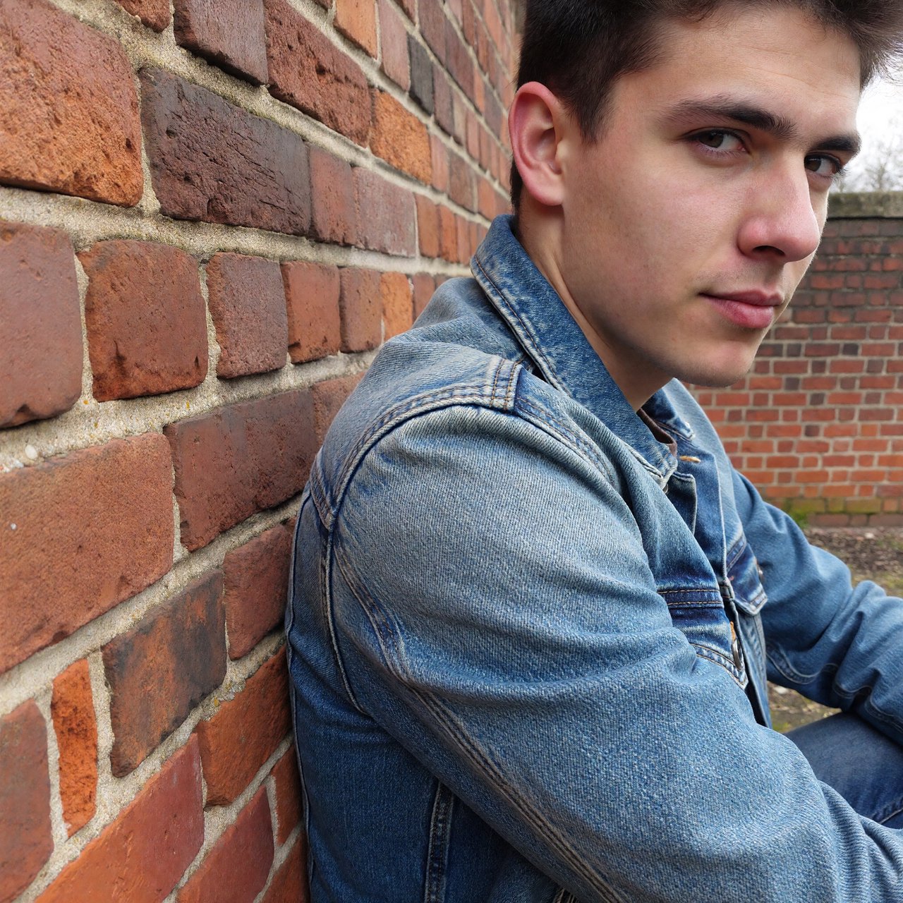 Environmental portrait of a young man in a denim jacket leaning on a red brick wall, overcast light, looking just past the camera. — ImagineArt 1.5 Pro