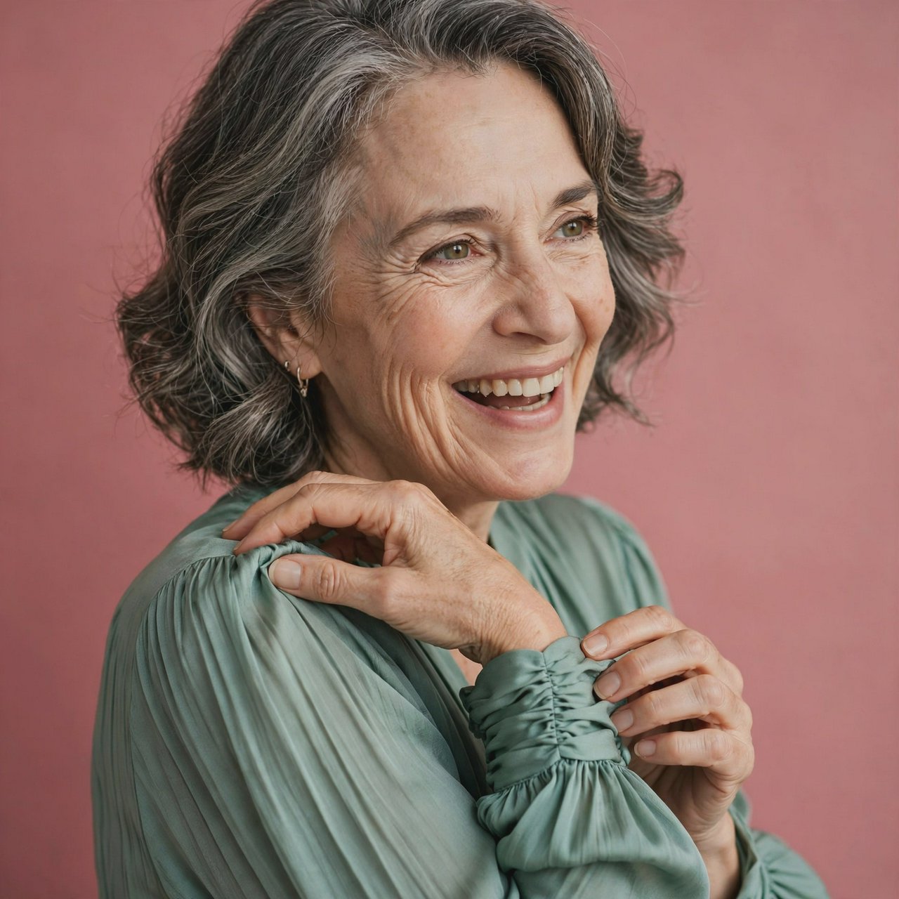 Portrait photograph of an adult woman in her early 60s wearing a softly pleated sea-green silk blouse, hands brushing its gathered sleeve fabric. Her amused, open expression stands against a matte rose backdrop, illuminated by broad, gentle studio light. — Muse Image 1.0