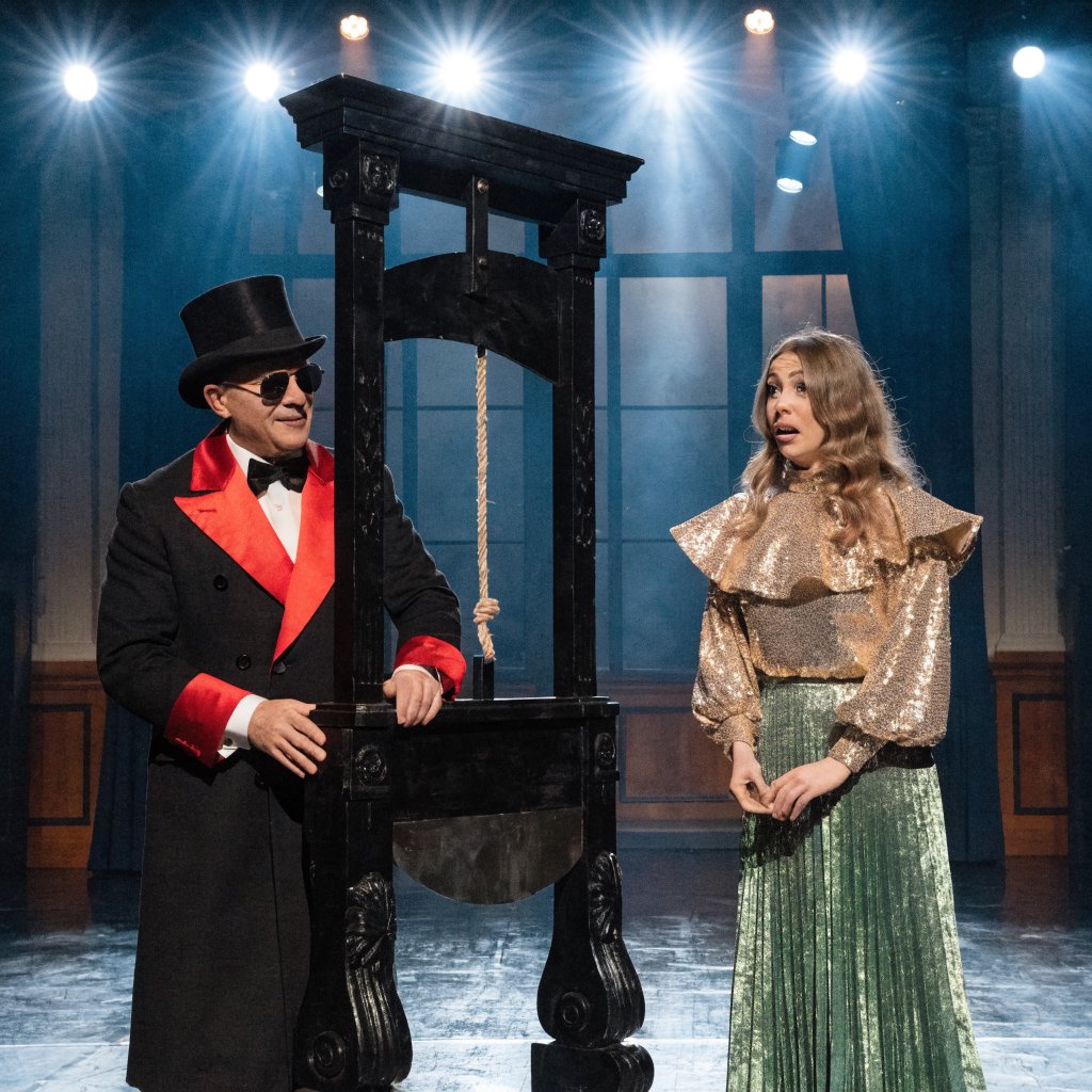 A magician's act on a well-lit stage. A well-dressed magician and glamorous adult female assistant have just unveiled the magician's guillotine. The adult female assistant fakes a worried look, knowing she will shortly participate in the illusion. — Recraft v4