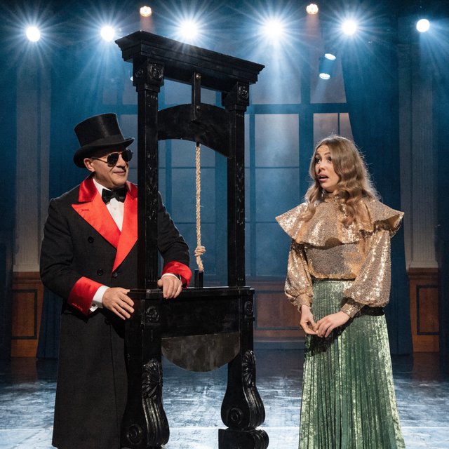A magician's act on a well-lit stage. A well-dressed magician and glamorous adult female assistant have just unveiled the magician's guillotine. The adult female assistant fakes a worried look, knowing she will shortly participate in the illusion. — Recraft v4