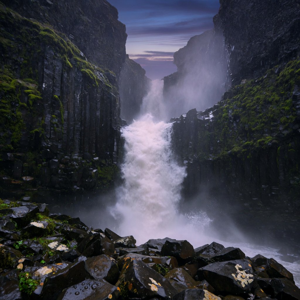 After a passing squall, a waterfall drops through a narrow basalt gorge at dusk, its white plume breaking against dark ledges; rain-beaded moss and sharp foreground stones lead toward cliffs fading behind drifting spray. — Grok Imagine Image Quality