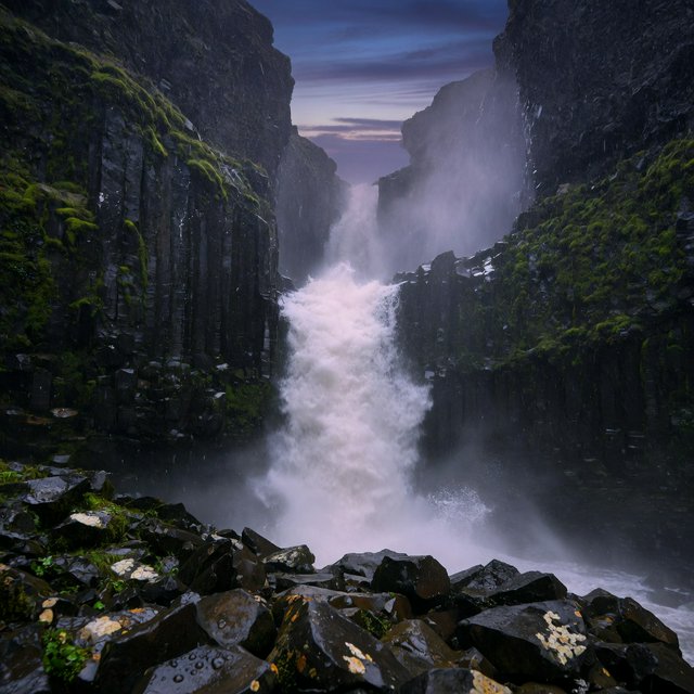 After a passing squall, a waterfall drops through a narrow basalt gorge at dusk, its white plume breaking against dark ledges; rain-beaded moss and sharp foreground stones lead toward cliffs fading behind drifting spray. — Grok Imagine Image Quality