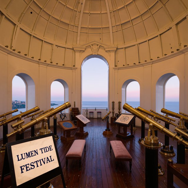 At a restored seaside observatory, a twilight meteor-watching event fills the domed hall with brass instruments, star charts, and velvet benches; "LUMEN TIDE FESTIVAL" is hand-printed in deep blue ink across a cream paper announcement beside the entrance. — Kling v3