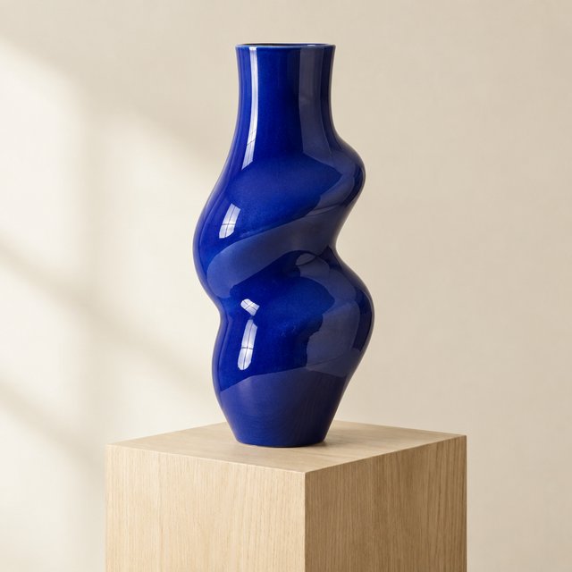 Commercial product photograph of an unbranded sculptural ceramic vase, formed from glossy cobalt porcelain, standing on a pale oak pedestal against a warm ivory background, illuminated by soft directional daylight and subtle fill. — GPT Image 2.5 Flare