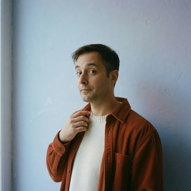 Portrait photograph of an adult man in his late 30s wearing a rust-colored corduroy overshirt over a cream knit, fingertips resting on its broad collar. His quietly curious gaze meets the camera against a pale blue plaster backdrop, softly lit by cool side-window light. — Grok Imagine Image Quality