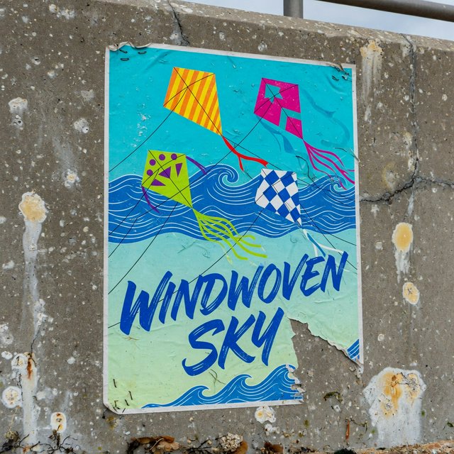 On a salt-stained seawall, a printed poster for a kite-making exhibition shows bright diamond kites sweeping above blue waves; “WINDWOVEN SKY” is screen-printed in sweeping cobalt script, with no other legible writing. — Model 6