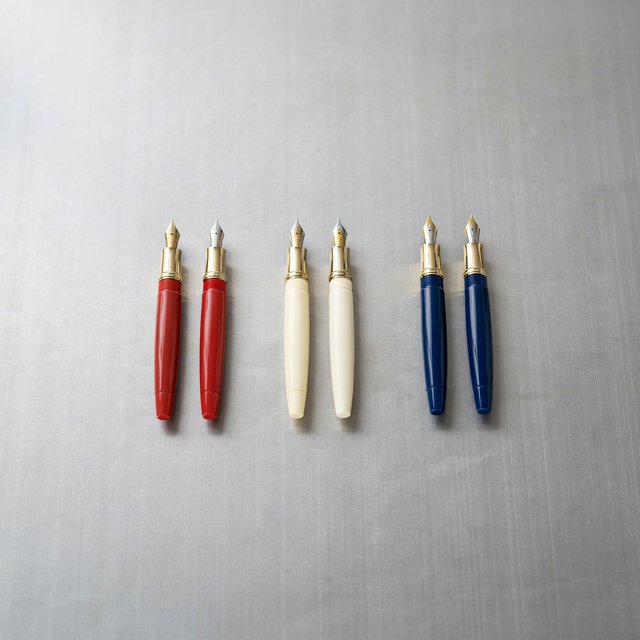 Across a brushed steel counter, exactly six fountain pens rest in three groups: two crimson pens on the left, two ivory pens in the center, and two cobalt pens on the right; nothing else. — Ideogram v4