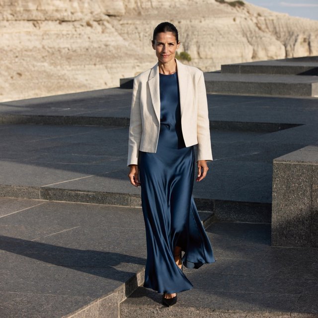 Across a basalt terrace, an adult woman in her early forties poses in a full-length fashion photograph, wearing a pleated indigo silk dress and cropped linen jacket, her composed half-smile lit by hard afternoon sunlight from one side against a pale limestone ridge. — ImagineArt 2