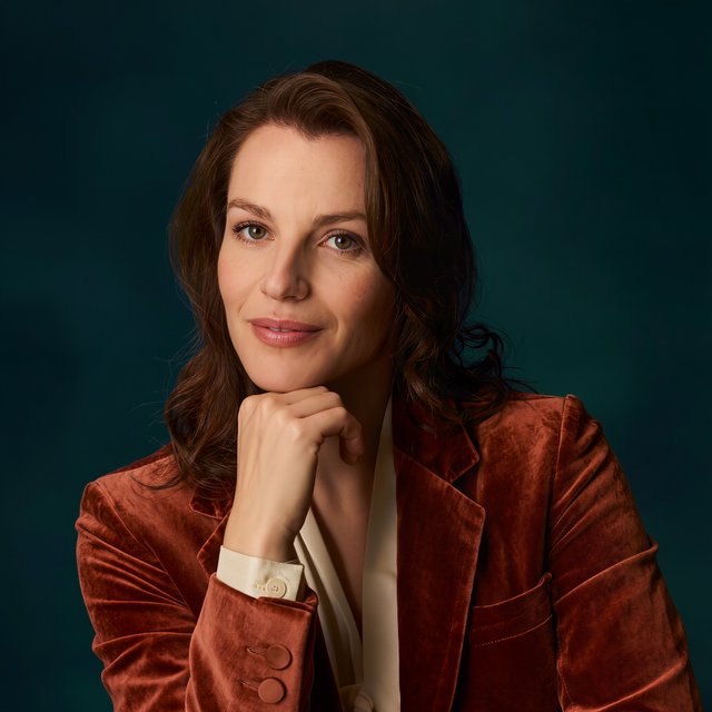 Portrait of an adult woman in her 30s wearing a rust-colored velvet jacket over an ivory blouse, one hand resting beneath her chin, thoughtful expression, deep teal studio backdrop, soft side lighting and gentle cheek highlights. — Kling v3