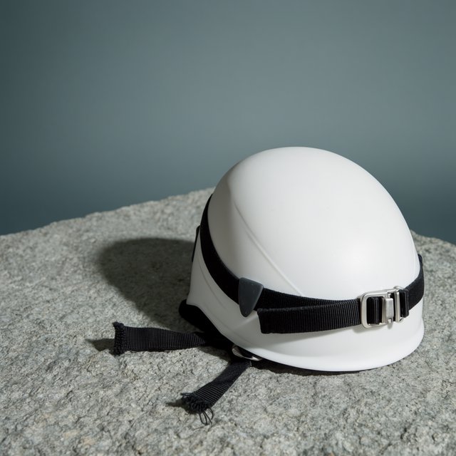 Against a muted slate backdrop, an unbranded climbing helmet with a matte recycled-polymer shell and woven nylon straps rests on rough granite; broad diffused key light and a cool rim light shape this commercial product image, with no lettering. — Krea 2 Large
