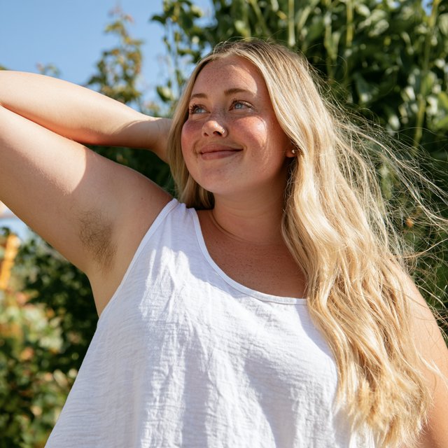 A vibrant medium shot of Cheyenne observing her armpit hair, her long blonde hair bouncing around her face, her blue eyes glimmering, and a soft smile playing on her lips. She is a 23-year-old adult with a very chubby body, a pointy nose, and thin blonde hairs on both forearms. She wears her signature white tank top and baggy shorts, standing against a backdrop of lush greenery and a sunny sky, with sunlight glimmering on her. — Midjourney V8.2