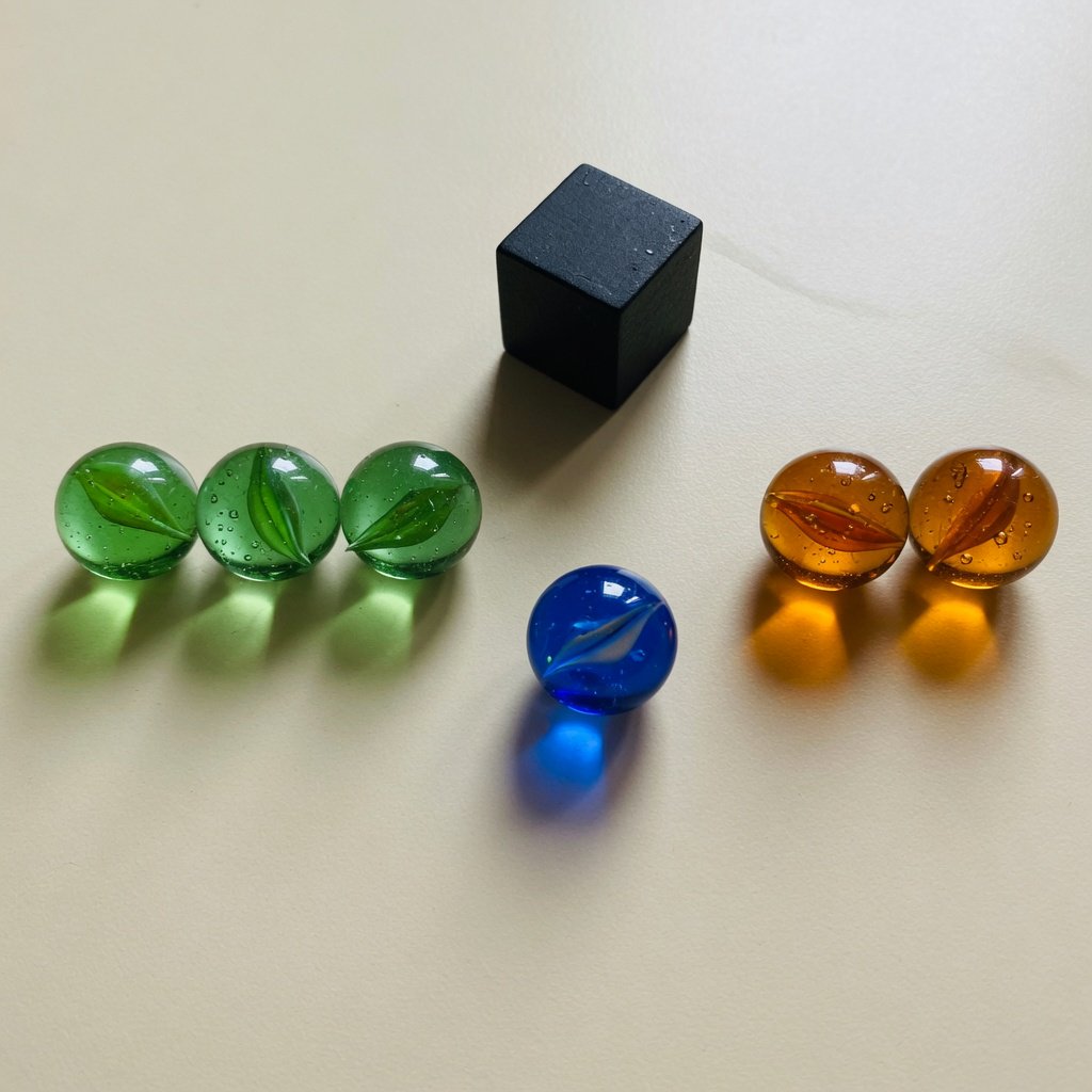 Exactly six glass marbles sit on a cream tabletop: three green marbles in a row on the left, two amber marbles in a row on the right, and one blue marble centered between the rows. A black cube sits above the blue marble, nothing else. — Grok Imagine Image Quality