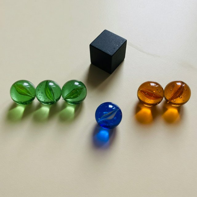 Exactly six glass marbles sit on a cream tabletop: three green marbles in a row on the left, two amber marbles in a row on the right, and one blue marble centered between the rows. A black cube sits above the blue marble, nothing else. — Grok Imagine Image Quality