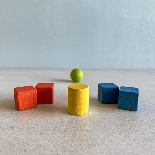 Exactly five wooden toy blocks sit on a pale gray table: two red cubes on the left, one yellow cylinder in the center, and two blue cubes on the right. A small green ball sits behind the cylinder, nothing else. — Midjourney V8.2