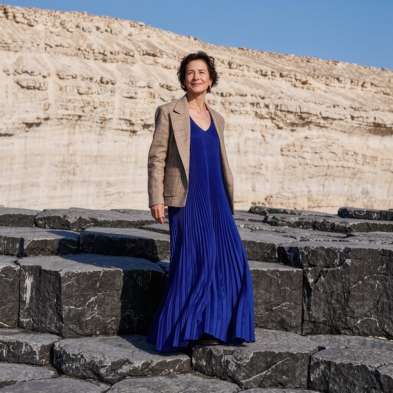 Across a basalt terrace, an adult woman in her early forties poses in a full-length fashion photograph, wearing a pleated indigo silk dress and cropped linen jacket, her composed half-smile lit by hard afternoon sunlight from one side against a pale limestone ridge. — Kling v3 Omni