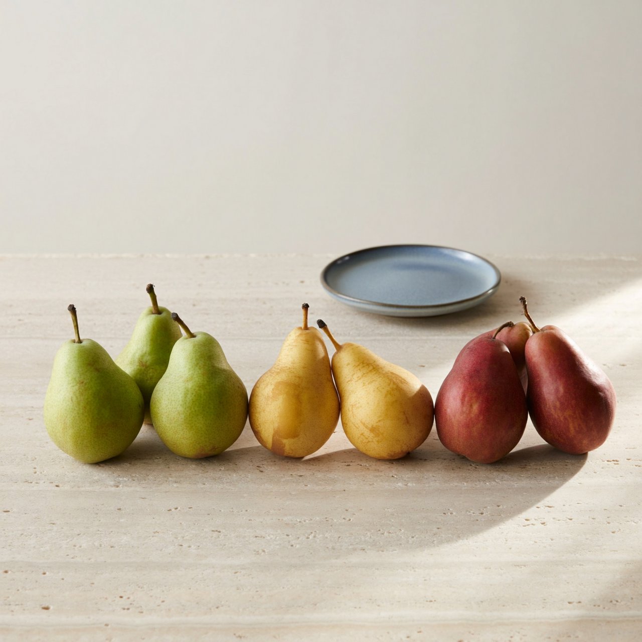 Exactly six ripe pears sit on a pale stone counter: two green pears on the left, two yellow pears in the center, and two red pears on the right. A small blue plate sits above the center pair, nothing else. — ImagineArt 2