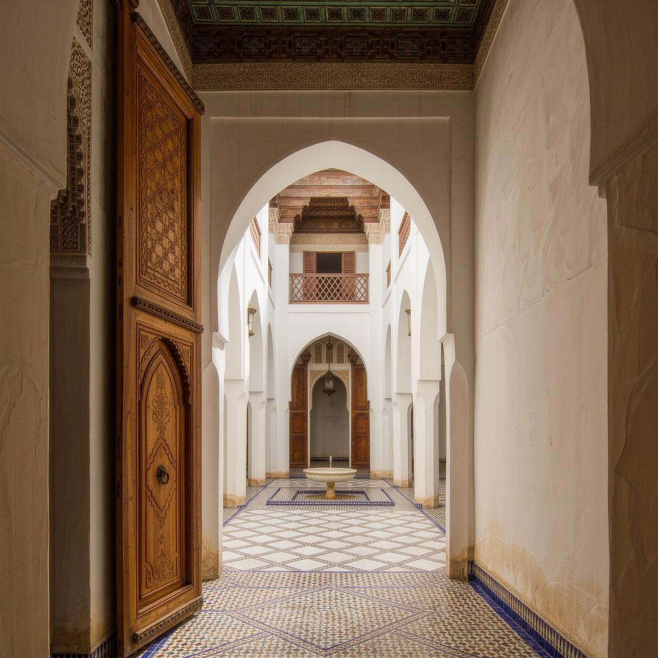 From a shaded courtyard entrance, Moorish Revival residences enclose a tiled fountain court, with cream stucco, carved cedar doors, and blue ceramic bands; late sunlight filters through arcades, revealing aligned walls and credible supports. — Kling v3 Omni