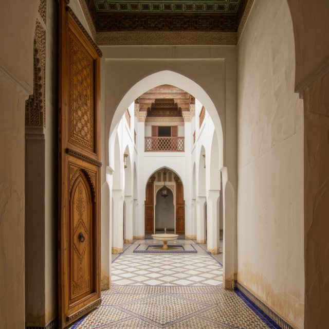 From a shaded courtyard entrance, Moorish Revival residences enclose a tiled fountain court, with cream stucco, carved cedar doors, and blue ceramic bands; late sunlight filters through arcades, revealing aligned walls and credible supports. — Kling v3 Omni