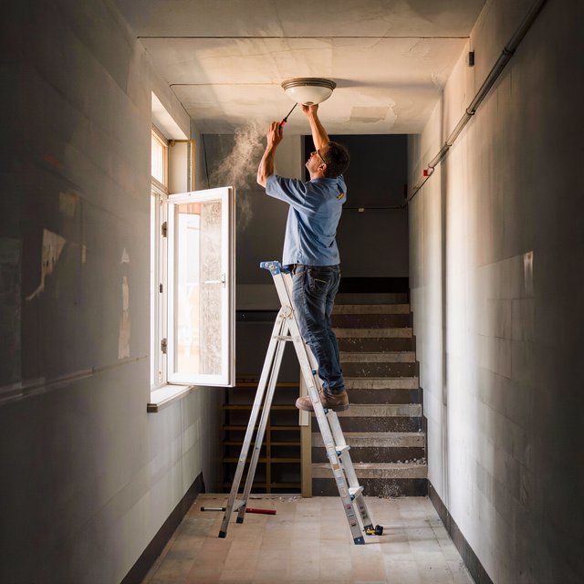 Candid documentary photograph of an adult building caretaker changing a ceiling light in a quiet apartment stairwell, pale afternoon window light, 35mm lens with moderate depth of field, plaster dust drifting from the fixture. — Kling v3