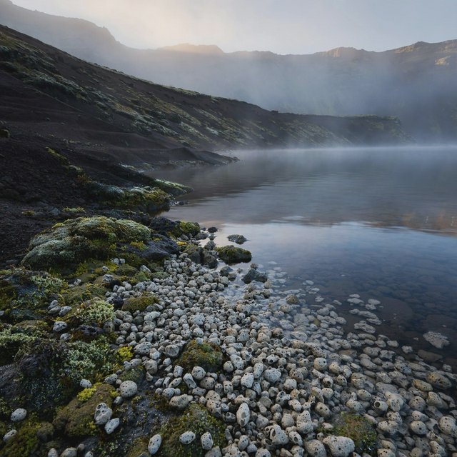 At first light, a rain-cleared volcanic caldera lake lies beneath thinning fog; pumice grains and damp lichen sharpen the near shore, while dark cinder slopes step back toward a distant rim. — Muse Image 1.0
