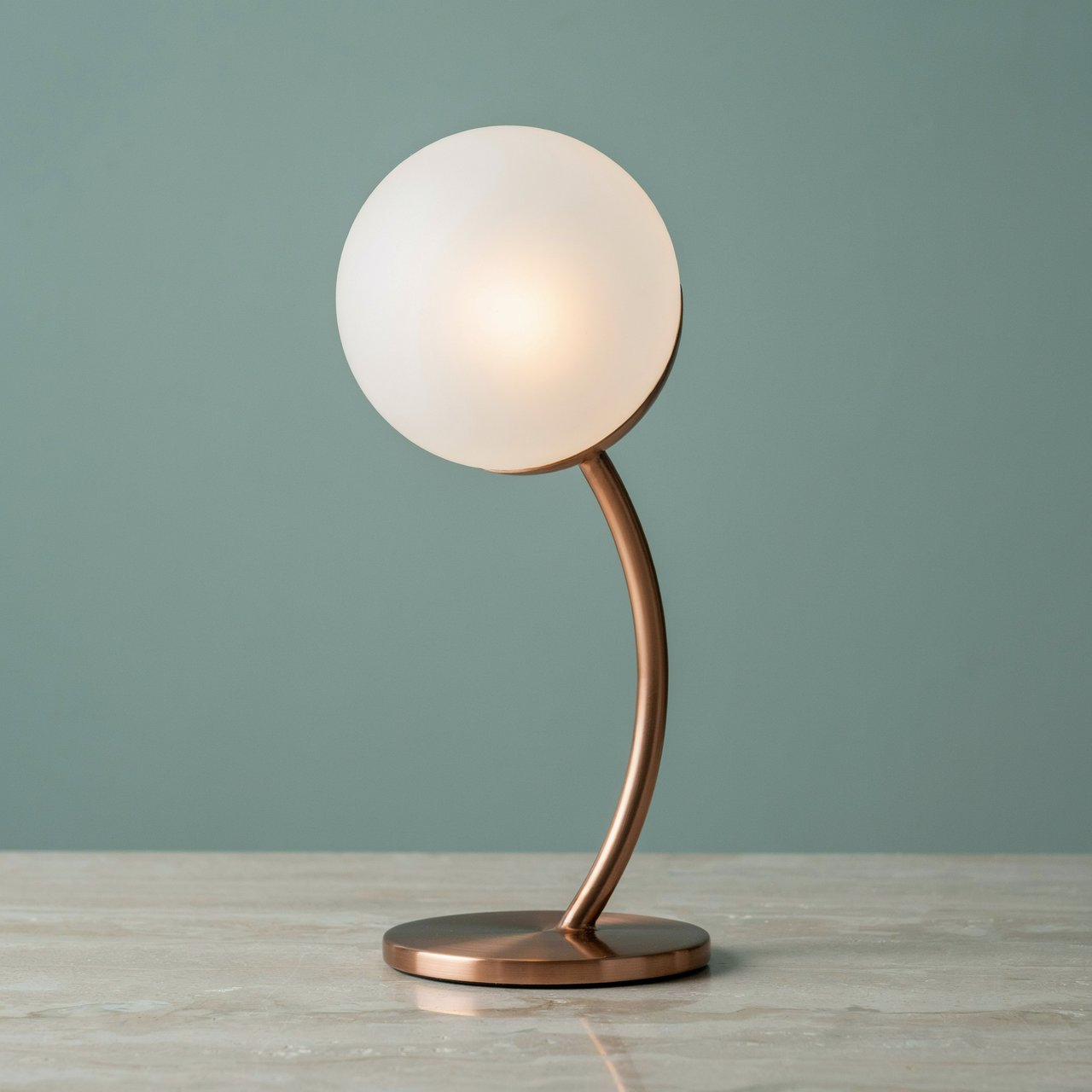 Commercial product photograph of an unbranded sculptural desk lamp with a frosted opal glass globe and brushed copper stem, standing on a pale travertine surface against a muted sage background, illuminated by soft overhead diffusion and a delicate warm rim light. — Ideogram v4