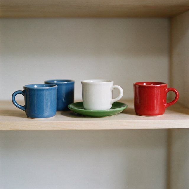 Exactly four ceramic mugs sit on a pale wooden shelf: two blue mugs on the left, one white mug in the center, and one red mug on the right. A green saucer is beneath the center mug, nothing else. — Seedream 5.0 Pro