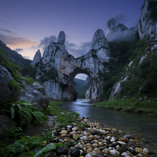 A limestone karst valley at blue hour after a light shower, towering pale cliffs rising above a clear river, low clouds snagging on pinnacles, slick ferns and rounded pebbles leading toward a distant stone arch. — Grok Imagine Image Quality