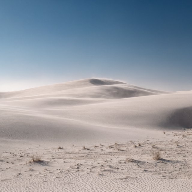 A pale gypsum dune field beneath a wind-brushed, cloudless midday sky, fine sand streaming over steep ridges; delicate ripple patterns and scattered salt-tolerant grasses sharpen the foreground as dunes soften toward a distant basin. — Krea 2 Large