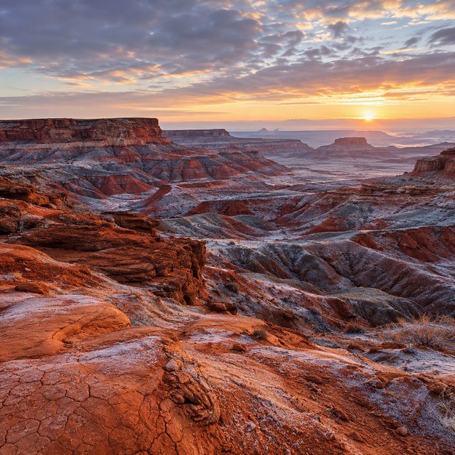 A red-sandstone badlands plateau beneath clearing winter clouds at sunrise, overnight frost silvering the gullies; cracked clay and wind-smoothed rock lead the eye across layered mesas fading into pale distance. — GPT Image 2.5 Flare