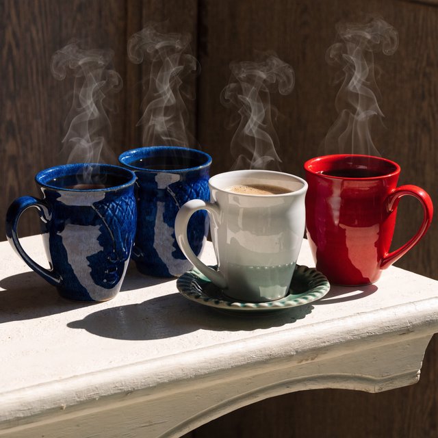 Exactly four ceramic mugs sit on a pale wooden shelf: two blue mugs on the left, one white mug in the center, and one red mug on the right. A green saucer is beneath the center mug, nothing else. — Recraft v4 Pro
