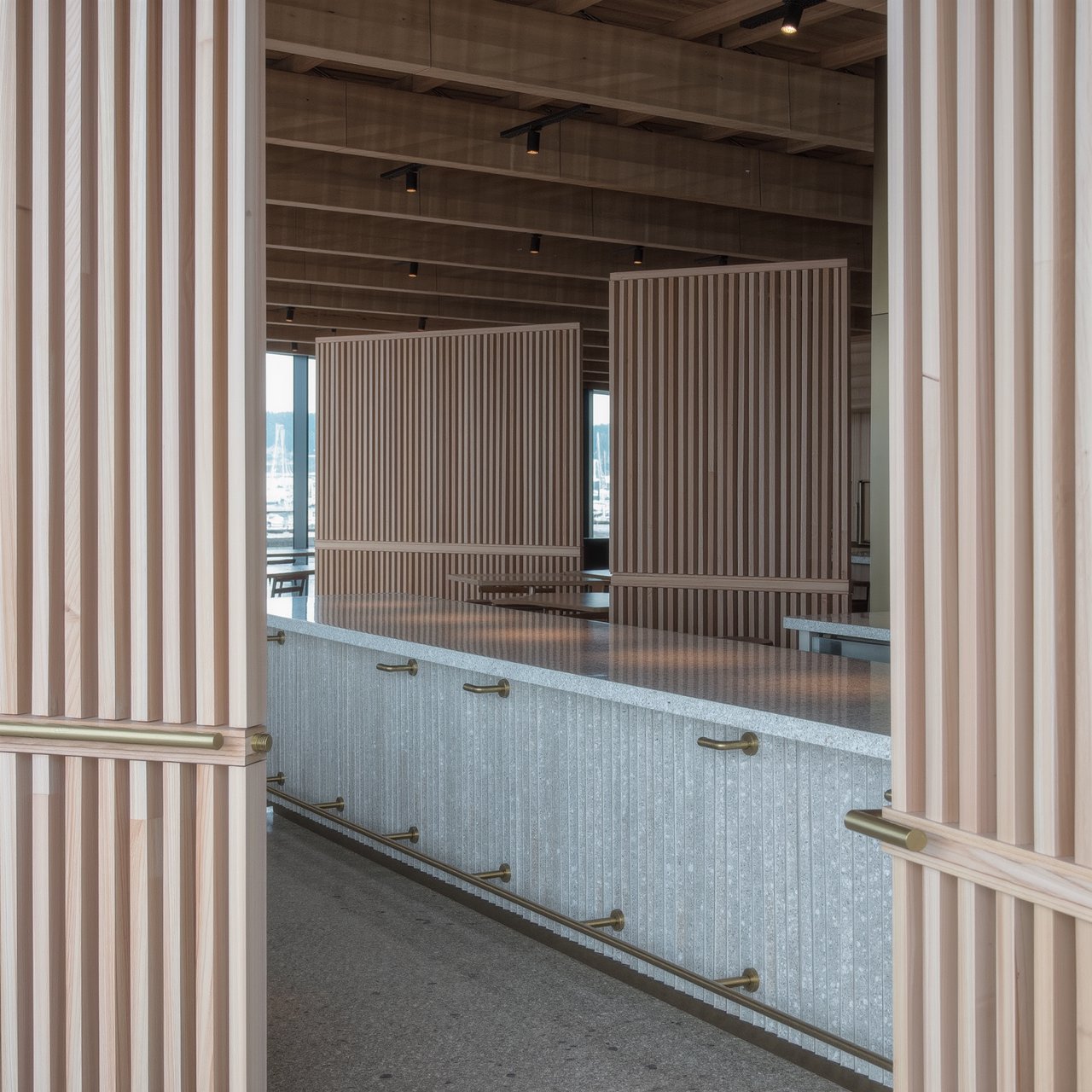 Inside an empty harbor café, restrained Danish modern design pairs ash slat screens, pale terrazzo, and brushed brass; overcast window light reaches the counter, viewed from the entrance, with aligned joinery and believable supports. — Kling v3 Omni