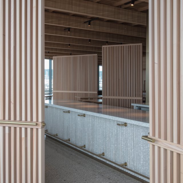 Inside an empty harbor café, restrained Danish modern design pairs ash slat screens, pale terrazzo, and brushed brass; overcast window light reaches the counter, viewed from the entrance, with aligned joinery and believable supports. — Model 10