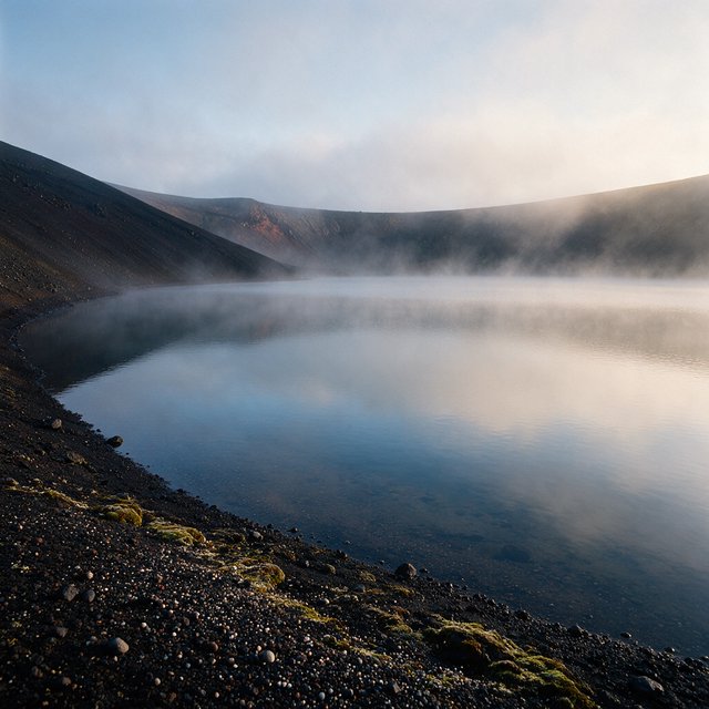 At first light, a rain-cleared volcanic caldera lake lies beneath thinning fog; pumice grains and damp lichen sharpen the near shore, while dark cinder slopes step back toward a distant rim. — Seedream 5.0 Pro
