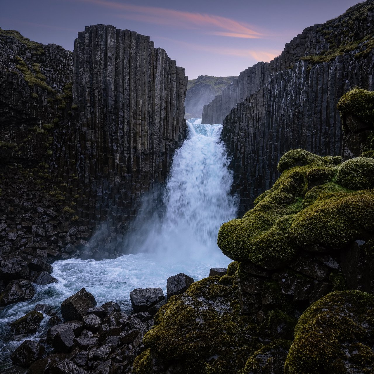 After a passing squall, a waterfall drops through a narrow basalt gorge at dusk, its white plume breaking against dark ledges; rain-beaded moss and sharp foreground stones lead toward cliffs fading behind drifting spray. — Kling v3 Omni