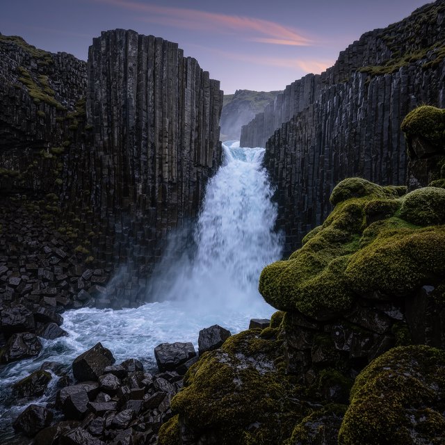 After a passing squall, a waterfall drops through a narrow basalt gorge at dusk, its white plume breaking against dark ledges; rain-beaded moss and sharp foreground stones lead toward cliffs fading behind drifting spray. — Kling v3 Omni