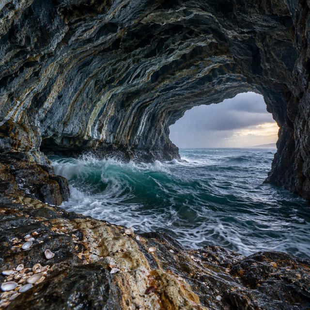Inside a blue-grey sea cave, incoming swells curl over ribbed limestone beneath a clearing rain squall in late afternoon; wet mineral bands and shell fragments sharpen the entrance ledge, while the cavern mouth frames hazy open water. — GPT Image 2.5 Flare