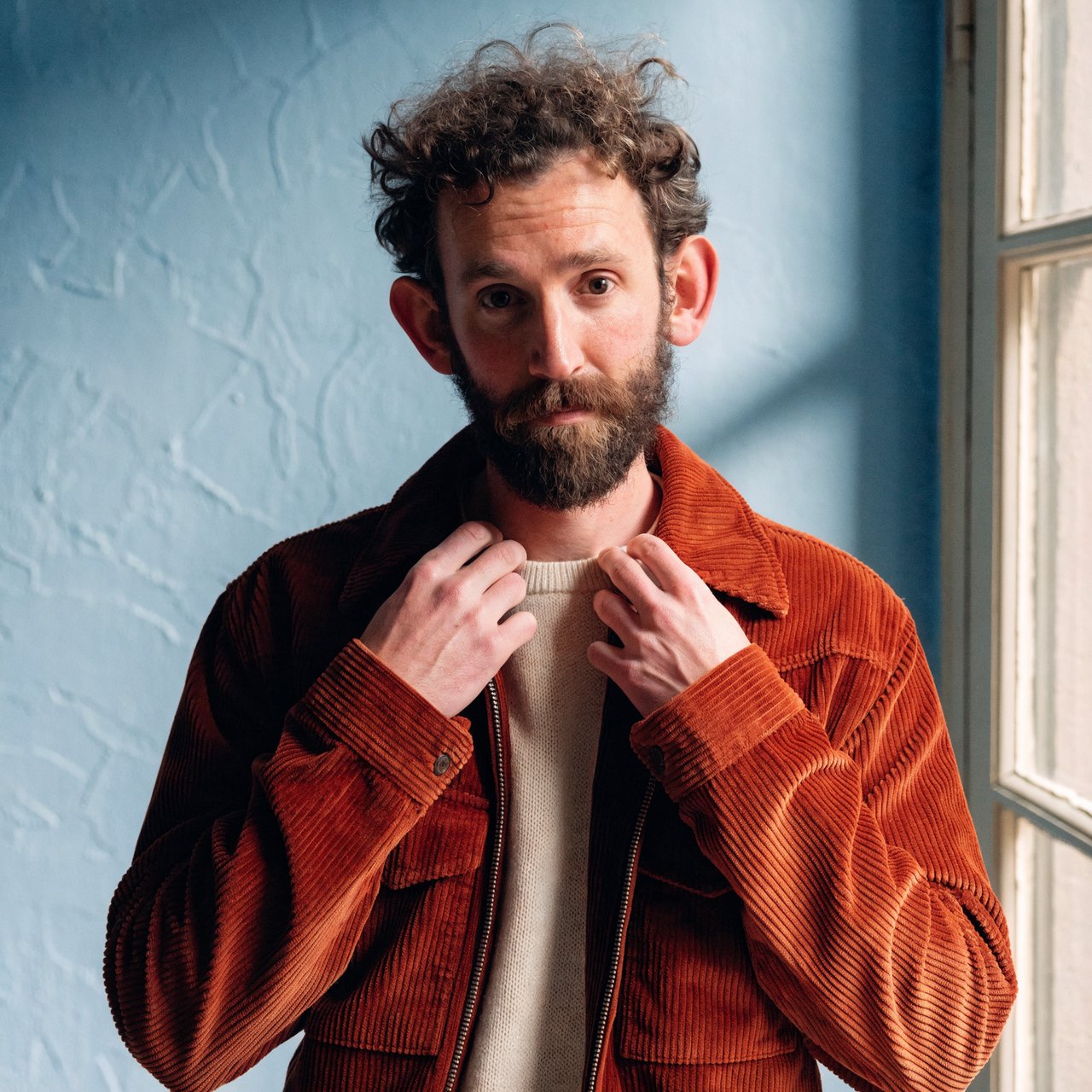 Portrait photograph of an adult man in his late 30s wearing a rust-colored corduroy overshirt over a cream knit, fingertips resting on its broad collar. His quietly curious gaze meets the camera against a pale blue plaster backdrop, softly lit by cool side-window light. — Recraft v4 Pro