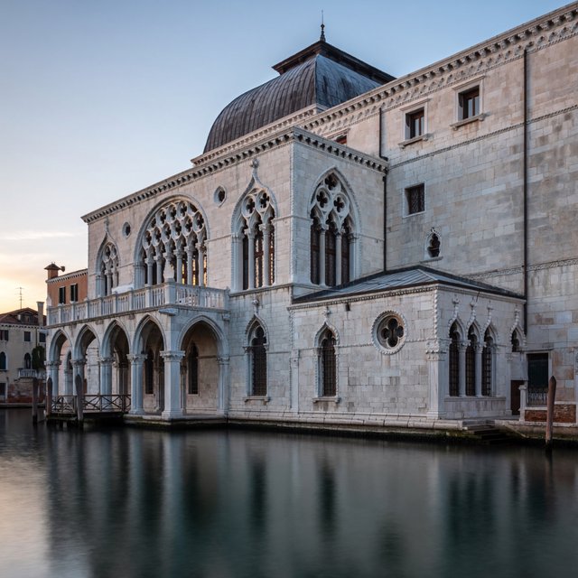 Exterior of a Venetian Gothic opera house beside a quiet canal, pale Istrian limestone, pointed arches, quatrefoil tracery, and a steep slate roof; cool morning light, viewed from across the water, crisp masonry lines and believable structure. — ImagineArt 2
