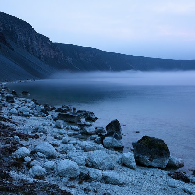 At first light, a rain-cleared volcanic caldera lake lies beneath thinning fog; pumice grains and damp lichen sharpen the near shore, while dark cinder slopes step back toward a distant rim. — Midjourney V8.2