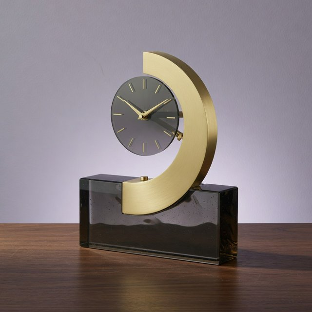 Commercial product photograph of an unbranded sculptural table clock made from satin brass and smoked glass, resting on a dark walnut surface against a muted lavender background, illuminated by a large diffused softbox from the upper left and a subtle rear rim light. — Grok Imagine Image Quality
