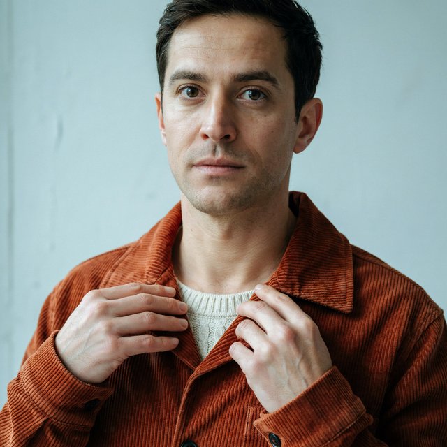 Portrait photograph of an adult man in his late 30s wearing a rust-colored corduroy overshirt over a cream knit, fingertips resting on its broad collar. His quietly curious gaze meets the camera against a pale blue plaster backdrop, softly lit by cool side-window light. — Ideogram v4.5