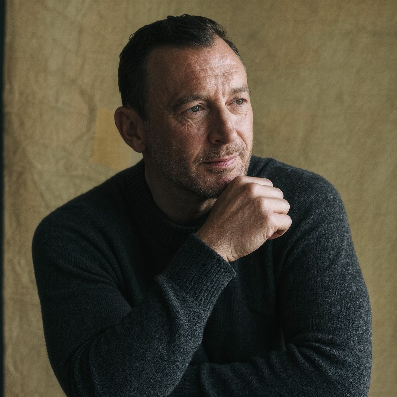 Editorial portrait of an adult man in his early 40s wearing a charcoal wool fisherman’s sweater, one hand loosely holding its ribbed cuff. His thoughtful, slightly tired expression rests against a muted ochre studio backdrop, shaped by gentle directional window light. — Kling v3 Omni