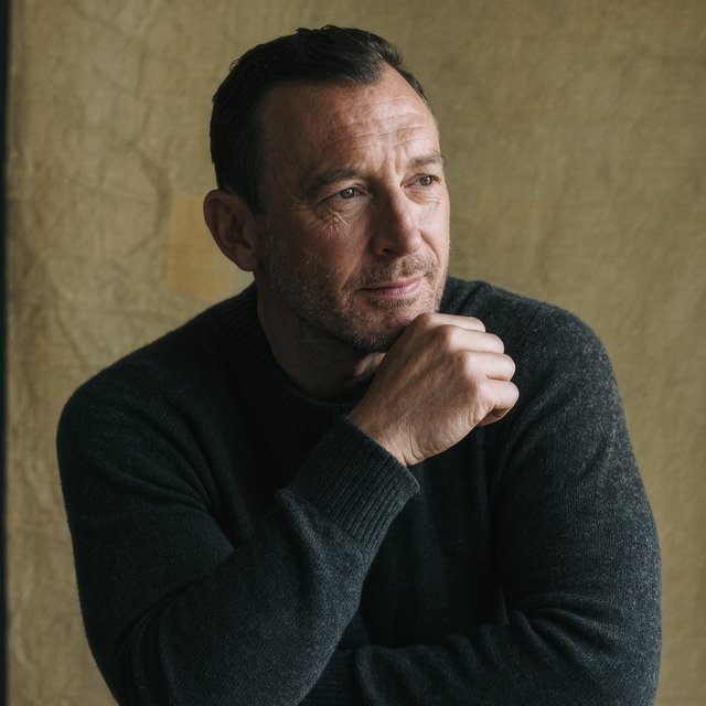 Editorial portrait of an adult man in his early 40s wearing a charcoal wool fisherman’s sweater, one hand loosely holding its ribbed cuff. His thoughtful, slightly tired expression rests against a muted ochre studio backdrop, shaped by gentle directional window light. — Kling v3 Omni