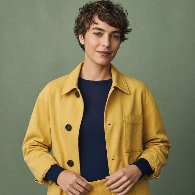 Fashion portrait of an adult nonbinary person in their late twenties wearing a structured saffron-yellow canvas jacket over a fine-gauge navy merino top, hands resting on the jacket hem. Their bright, self-assured expression contrasts with a softly lit sage-green backdrop. — Grok Imagine Image 2