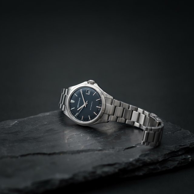 Product shot of a minimalist steel wristwatch with a navy dial on dark slate, single softbox from the upper left, subtle reflection. — Ideogram v4