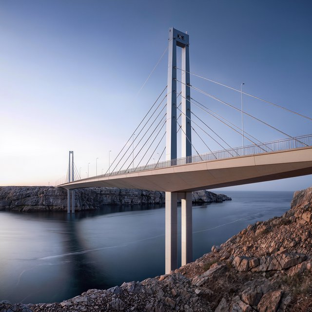 Across a sheltered coastal inlet, a contemporary high-tech cable-stayed bridge spans between granite shores, its slender steel pylons and pale concrete deck catching cool sunrise light; viewed from a rocky bank, straight lines and credible engineering define the scene. — Kling v3