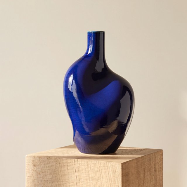 Commercial product photograph of an unbranded sculptural ceramic vase, formed from glossy cobalt porcelain, standing on a pale oak pedestal against a warm ivory background, illuminated by soft directional daylight and subtle fill. — ImagineArt 2